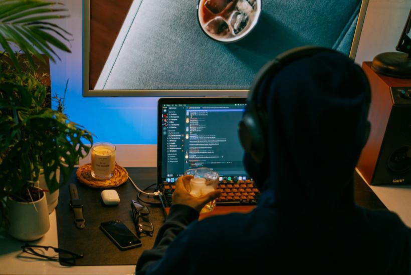 Хакер в капюшоне сидит перед монитором с кодом